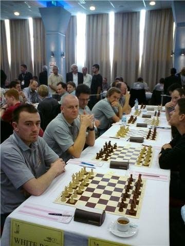 A row of players seated along a team-match table, boards and clocks set out and a 'White Rose' banner on the table edge; I'm at the near end, mid-game.